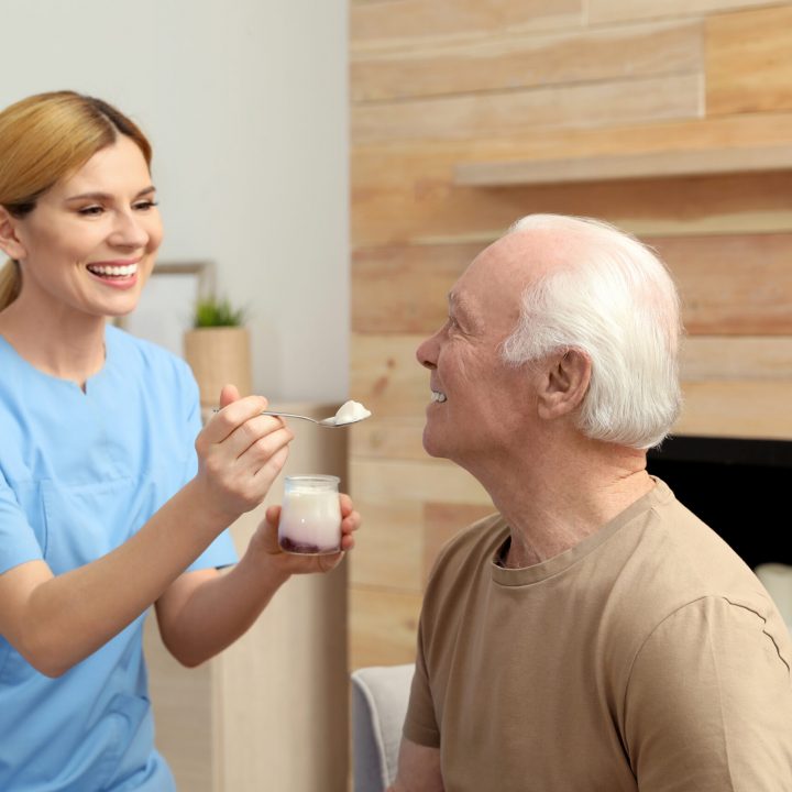 Nurse feeding elderly man with yogurt indoors. Assisting senior people